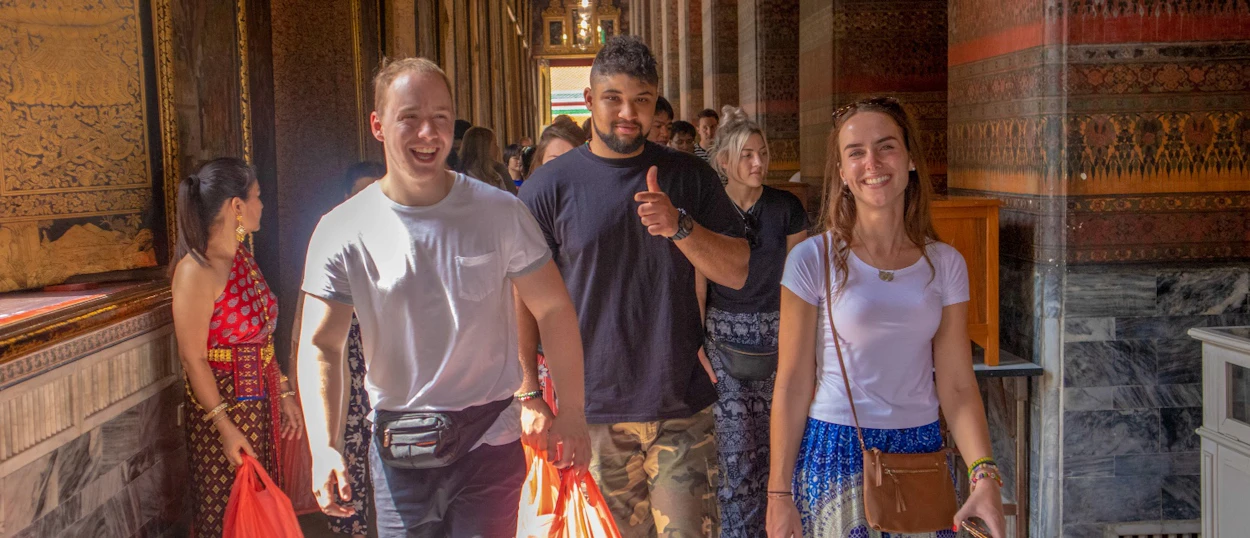 Happy tourists explore an ornate temple corridor in Bangkok, smiling and giving a thumbs up.