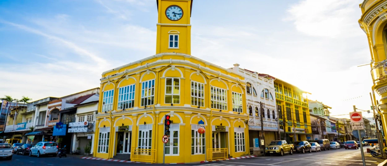 A vibrant yellow clock tower building anchors a sunny street lined with colorful architecture in Phuket.
