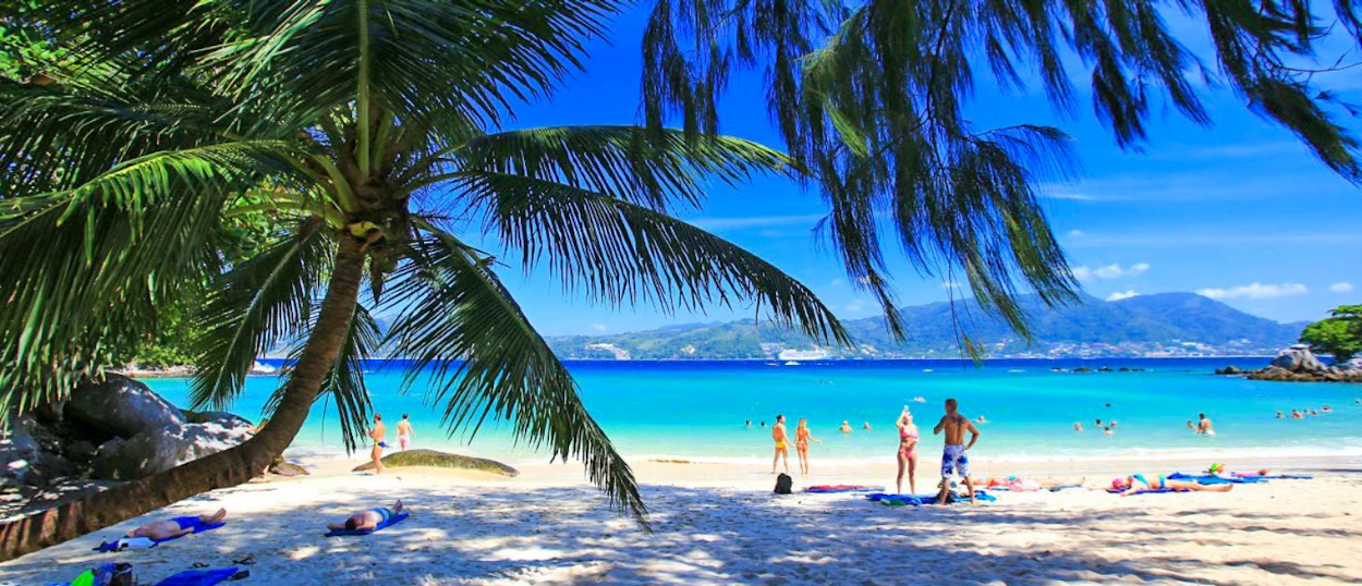 People relax on a sunny white sand beach with turquoise water and palm trees in Phuket.