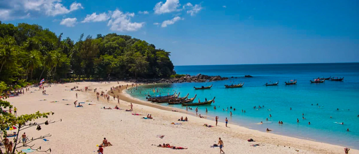 People enjoy the sunny white sand beach and turquoise water with longtail boats in Phuket.