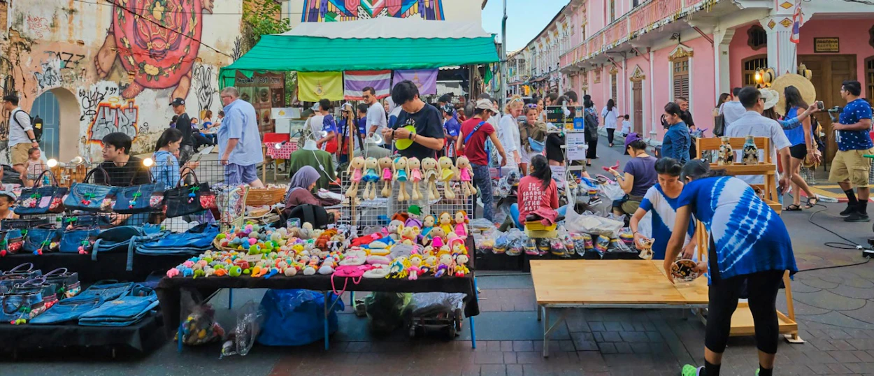 A bustling street market in Phuket Old Town with people browsing colorful stalls selling various goods.