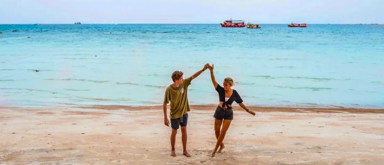 A happy couple dances playfully on Sairee Beach, Koh Tao, with blue ocean and boats.