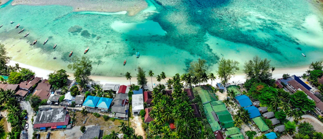 Aerial view of Koh Phangan's turquoise waters, sandy beach, and coastal buildings with anchored boats.