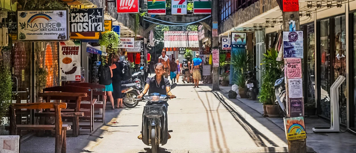 A person rides a scooter down a sunny, bustling street lined with shops and people in Koh Phangan.