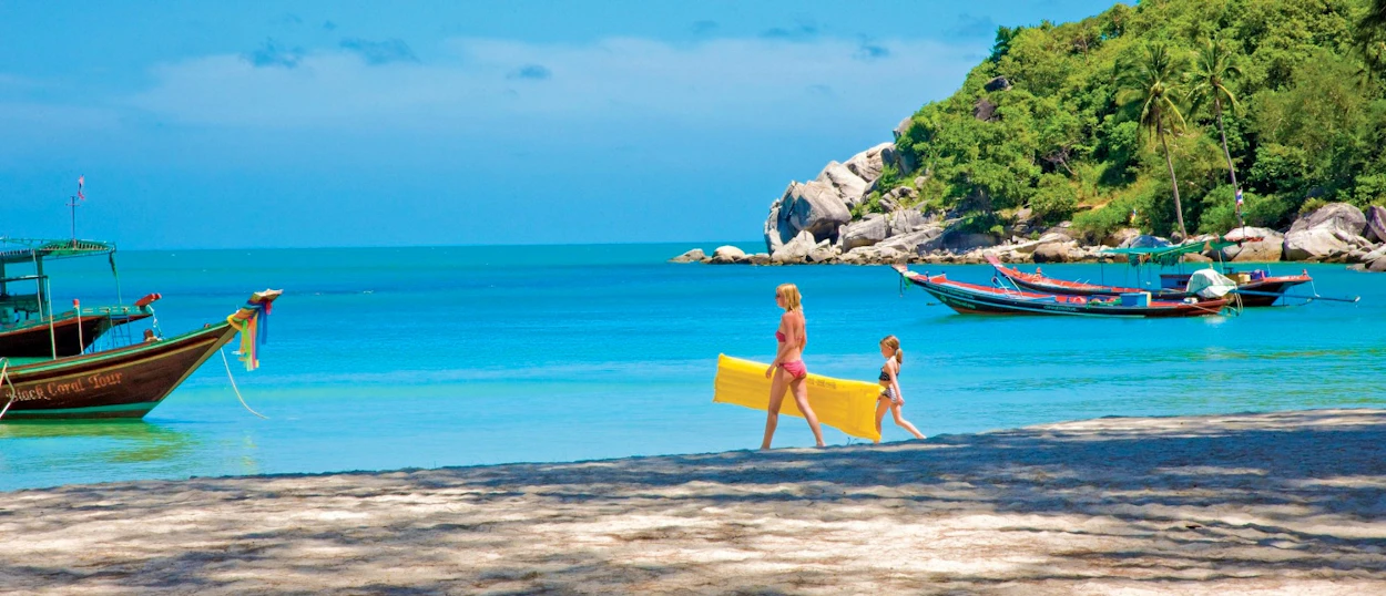 Mother and child walk on a sunny Koh Phangan beach with longtail boats in blue water.