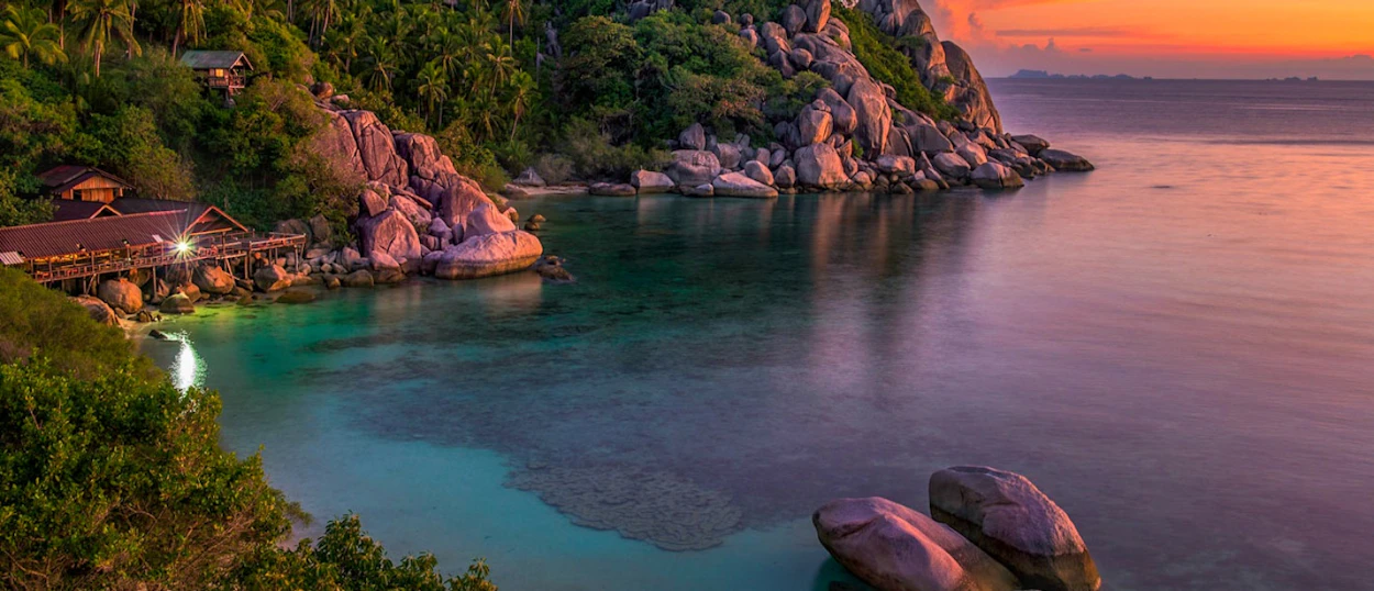 Tranquil tropical bay on Koh Phangan at sunset, with clear water, rocky shores, and a lit building.
