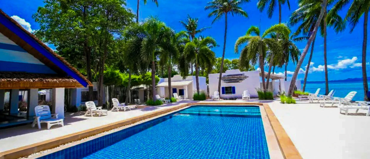 A vibrant blue swimming pool surrounded by palm trees and white buildings near the ocean in Koh Phangan.