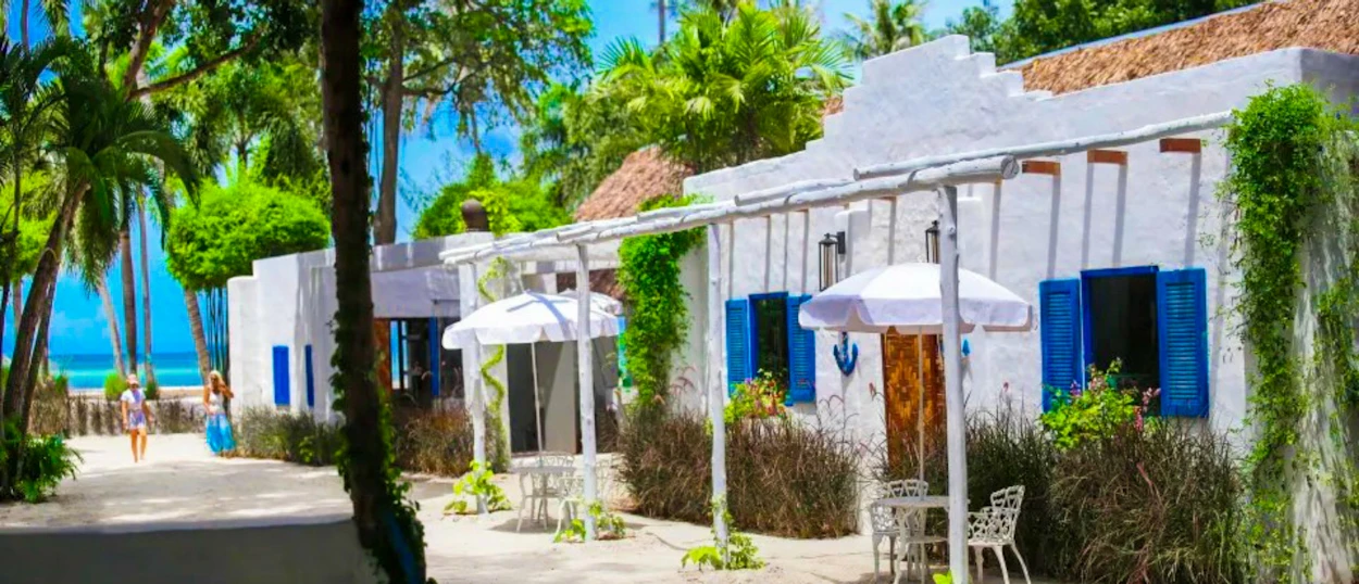 Two people walk past white bungalows with blue shutters towards a tropical beach in Koh Phangan.