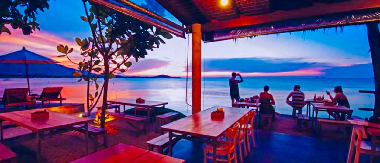 Tourists relax at a vibrant beachfront bar in Koh Samui during a beautiful sunset.