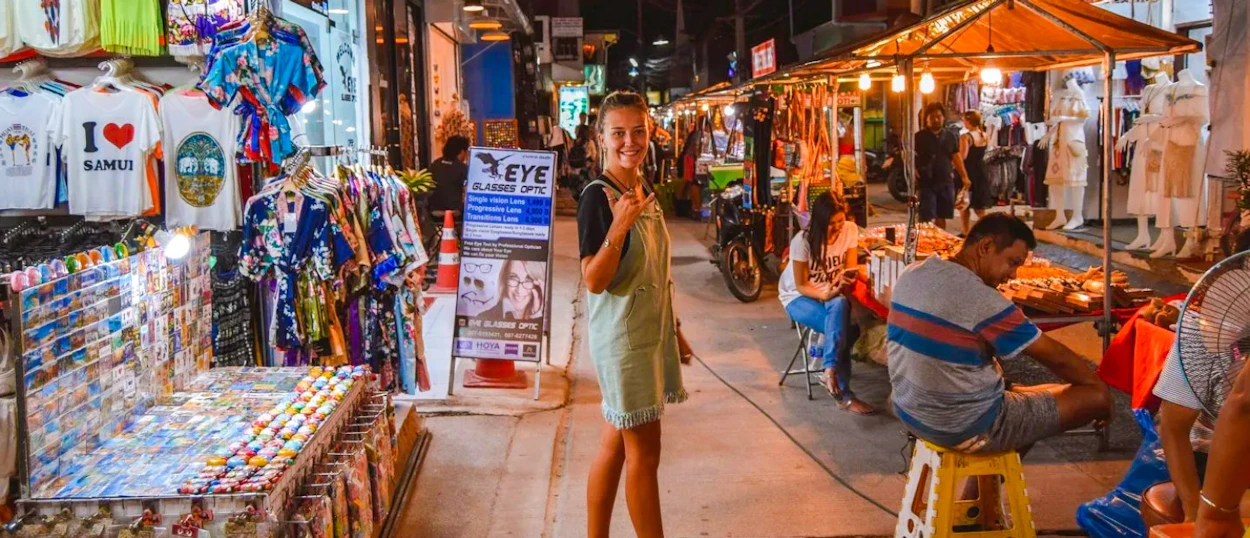 A smiling woman stands in a vibrant night market street with colorful stalls in Koh Samui.