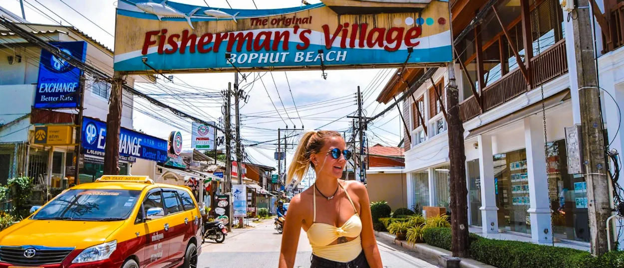 A smiling woman in sunglasses walks through the vibrant Fisherman's Village, Bophut Beach, Koh Samui.