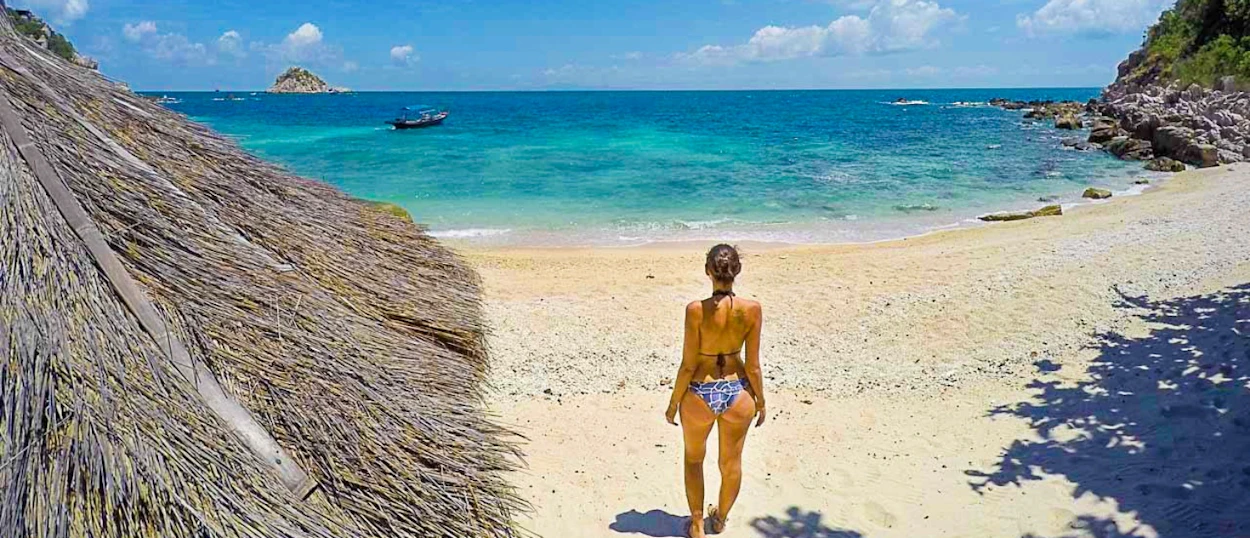 A woman walks on a white sand beach towards the clear turquoise ocean on Koh Tao.