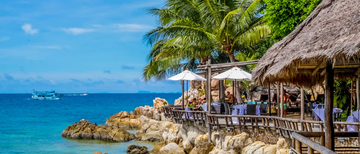 A beautiful thatched-roof restaurant on a wooden deck overlooks the turquoise ocean in Thailand.