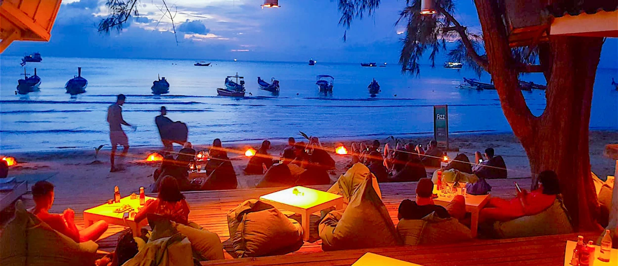 People relax at a lit beach bar with longtail boats on the sea at dusk in Koh Tao.