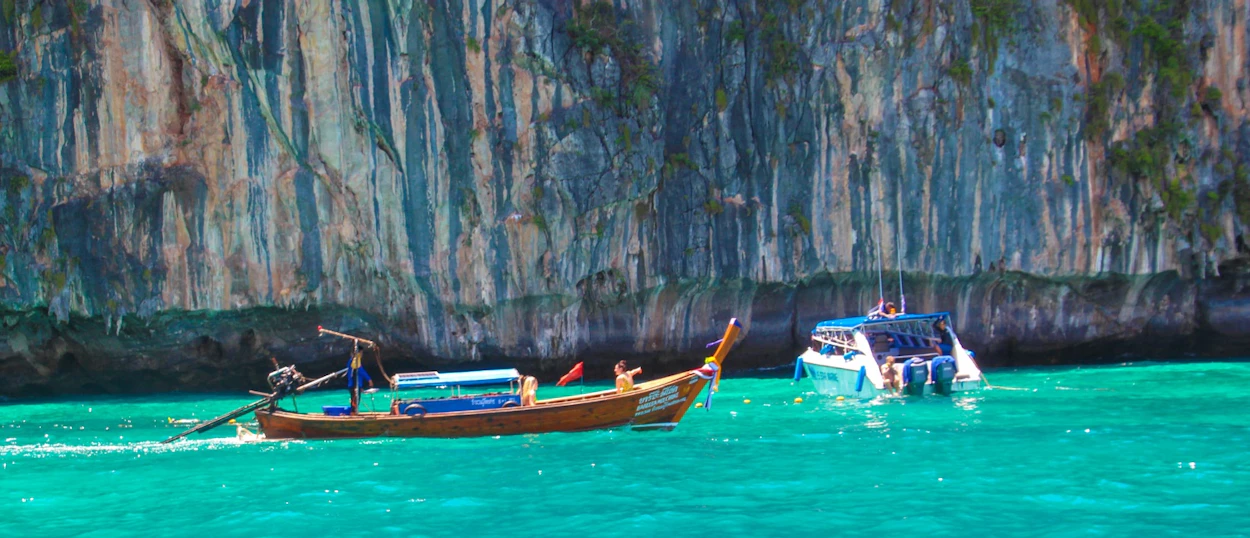 A traditional longtail boat and a speedboat navigate turquoise waters past dramatic limestone cliffs in Phuket.