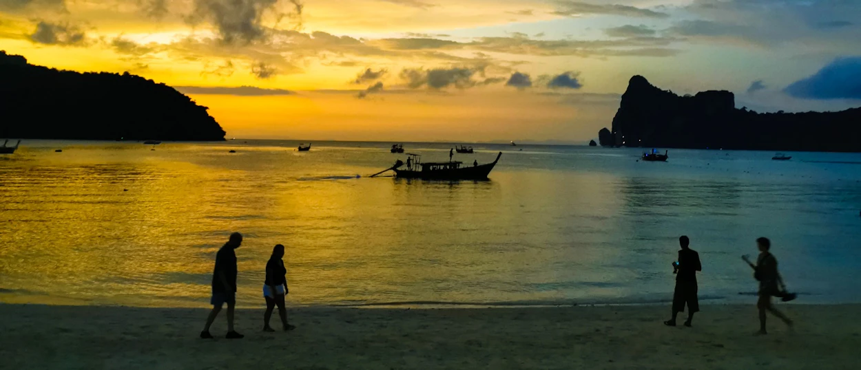 People stroll along a sandy beach at sunset with longtail boats and islands in Phuket.