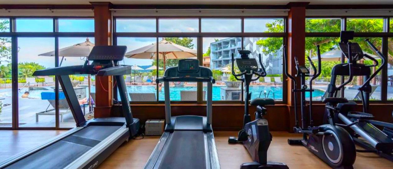 A hotel gym with treadmills and ellipticals overlooks a resort swimming pool in Phuket.