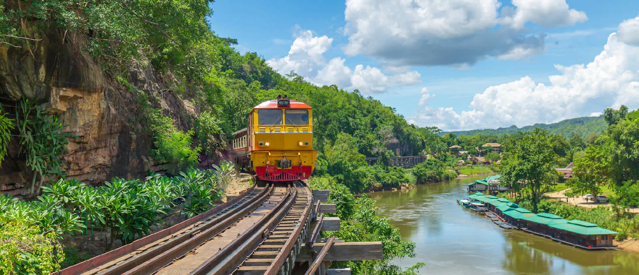 A yellow and red train travels on tracks alongside a lush green cliff above a calm river.