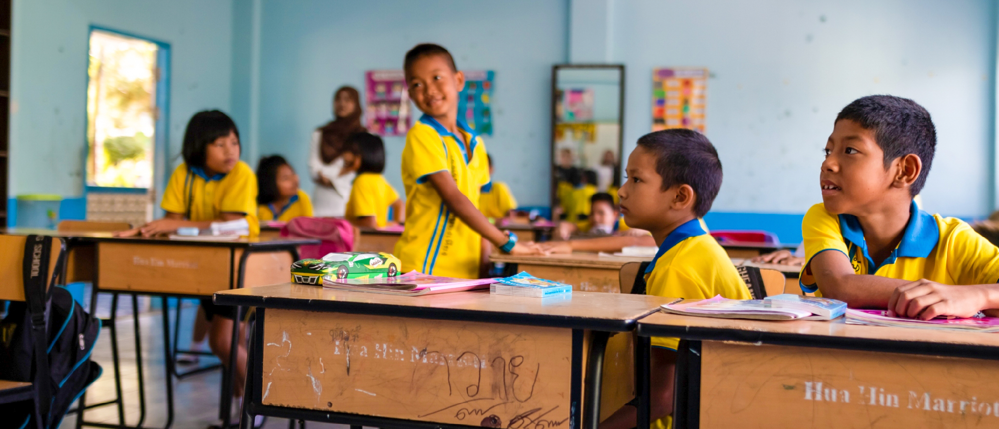 Smiling Thai students in yellow uniforms learning in a bright classroom, one boy standing.