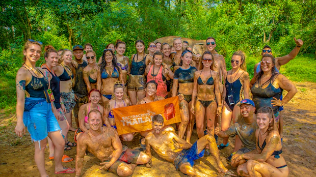A joyful group of young backpackers covered in mud, posing in a lush jungle in Thailand.