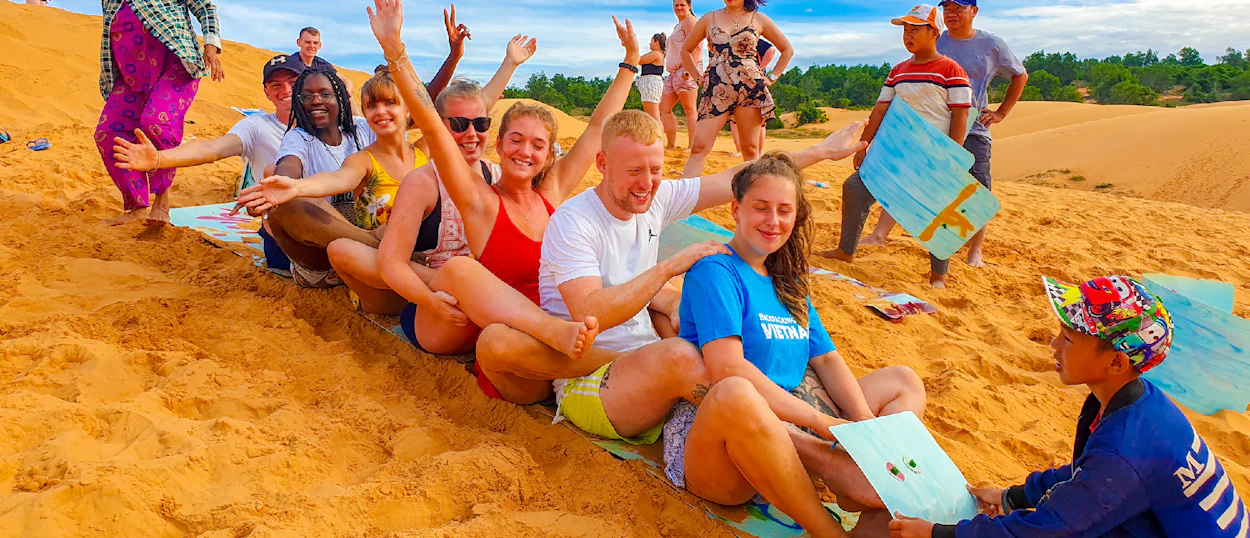 Joyful backpackers sit together on vibrant sand dunes, waving and ready for fun in Mui Ne, Vietnam.