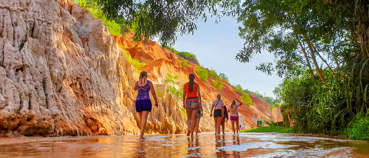 Backpackers walk through the shallow Fairy Stream in Mui Ne, surrounded by vibrant red and white canyon walls.