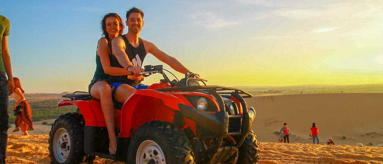 Smiling couple on a red ATV exploring the vast sand dunes of Mui Ne at sunrise.