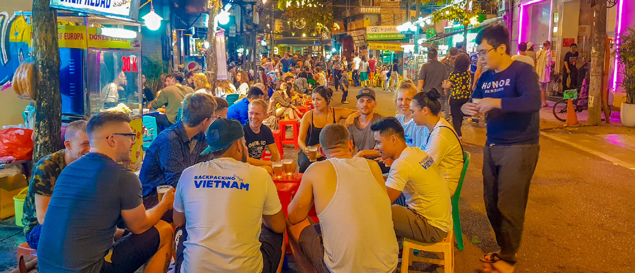 Backpackers gather around street tables, enjoying drinks amidst the vibrant, neon-lit nightlife of Hanoi.