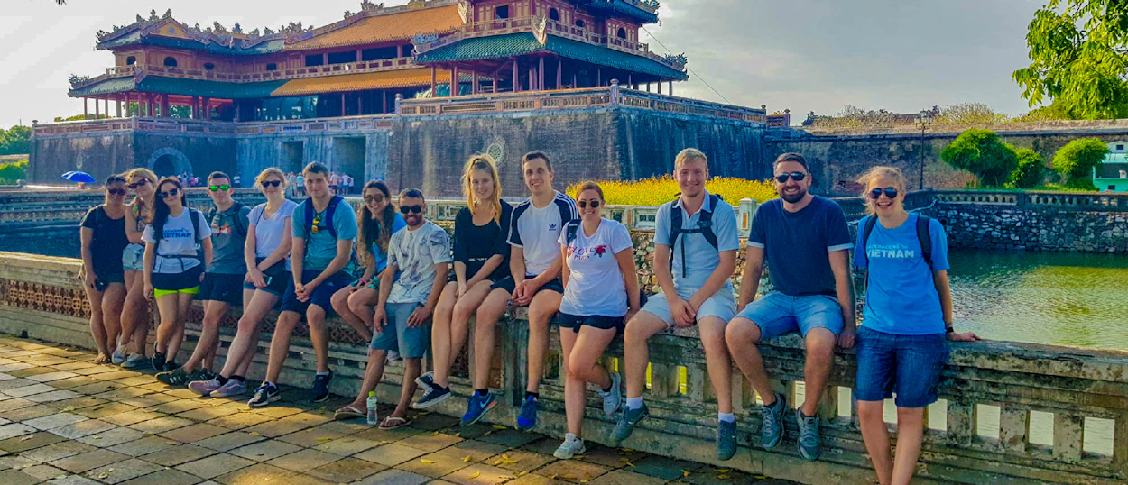 Smiling backpackers pose on a stone wall with the grand Imperial City of Hue in the background.