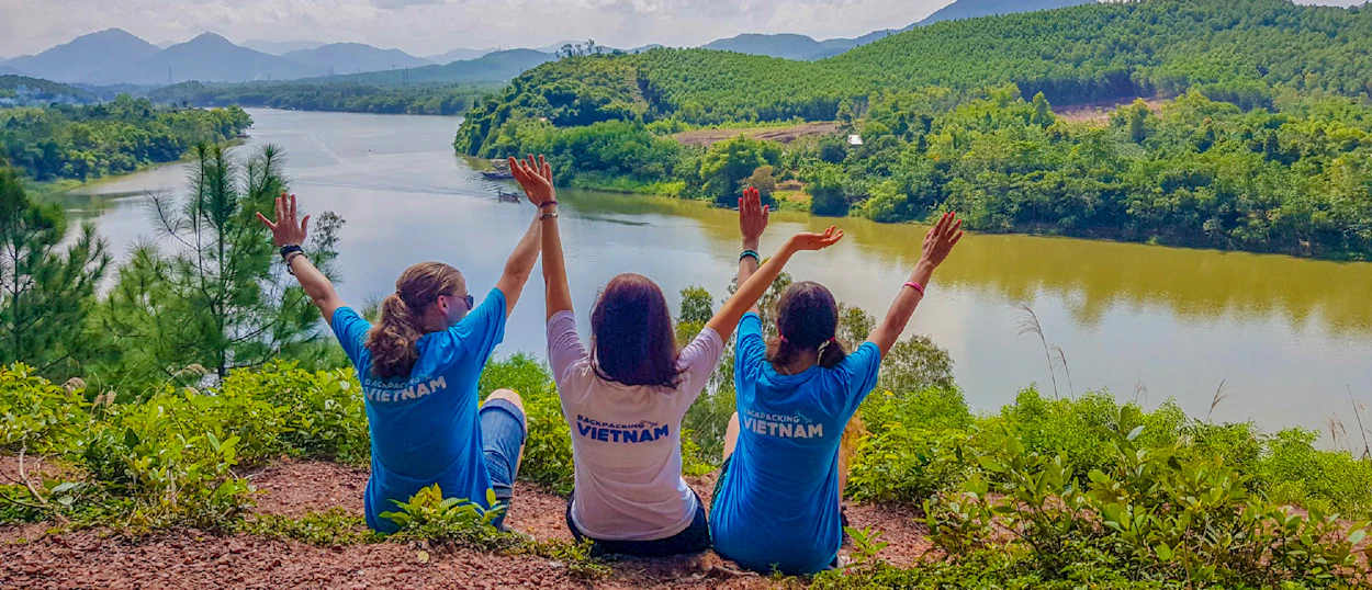 Three women with raised arms enjoy a panoramic view of a river and green hills in Vietnam.