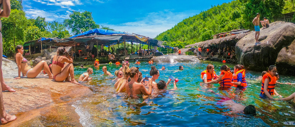 Backpackers swim and relax in a vibrant natural spring, surrounded by lush green hills in Vietnam.