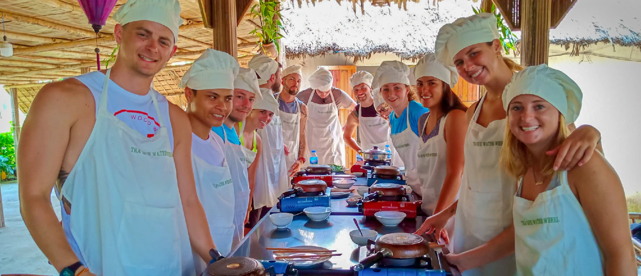 Smiling travelers in chef hats cooking together at an outdoor class in Hoi An, Vietnam.