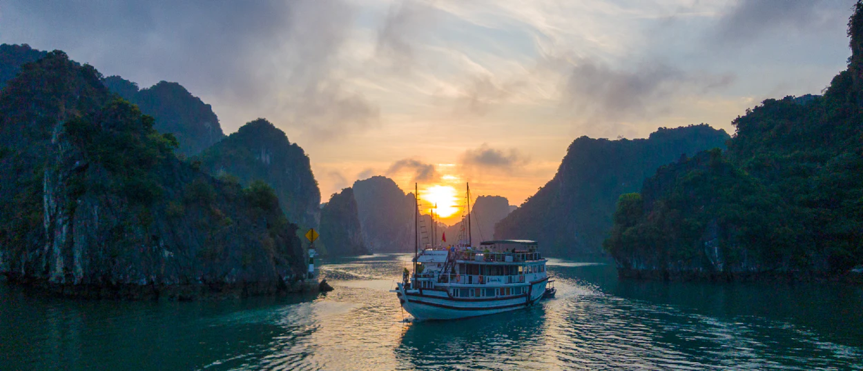 A boat cruises through the stunning limestone karsts of Halong Bay at sunset.