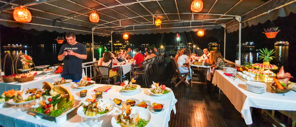 Travelers enjoy a lavish buffet dinner on a cruise boat deck at night in Halong Bay.