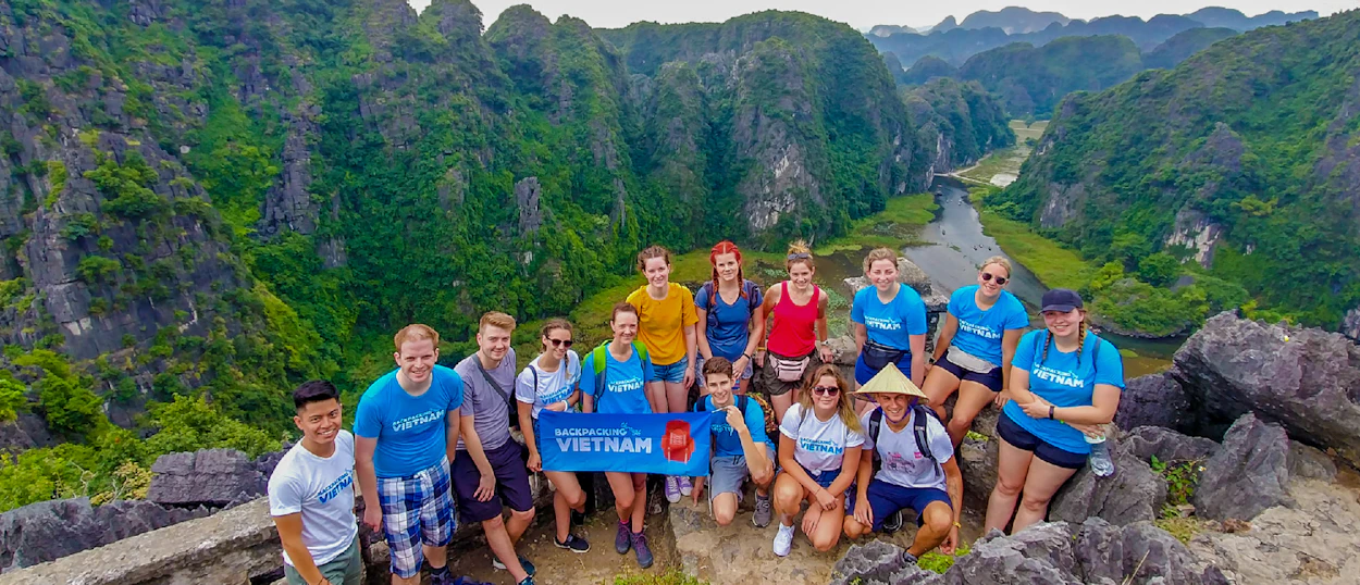 A group of happy backpackers pose on a mountain peak, overlooking the scenic Tam Coc river valley in Vietnam.