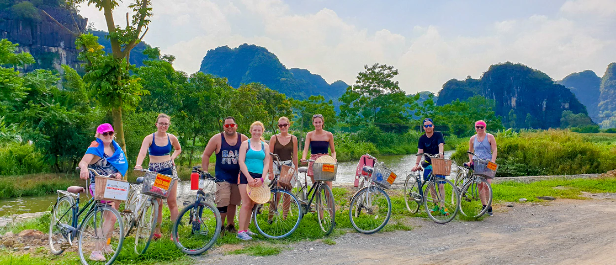 Eight smiling travelers with bicycles stand by a river, surrounded by dramatic limestone karsts in Tam Coc, Vietnam.