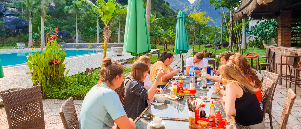 Travelers enjoy an outdoor meal by a resort pool, surrounded by tropical plants and Phong Nha's karst mountains.