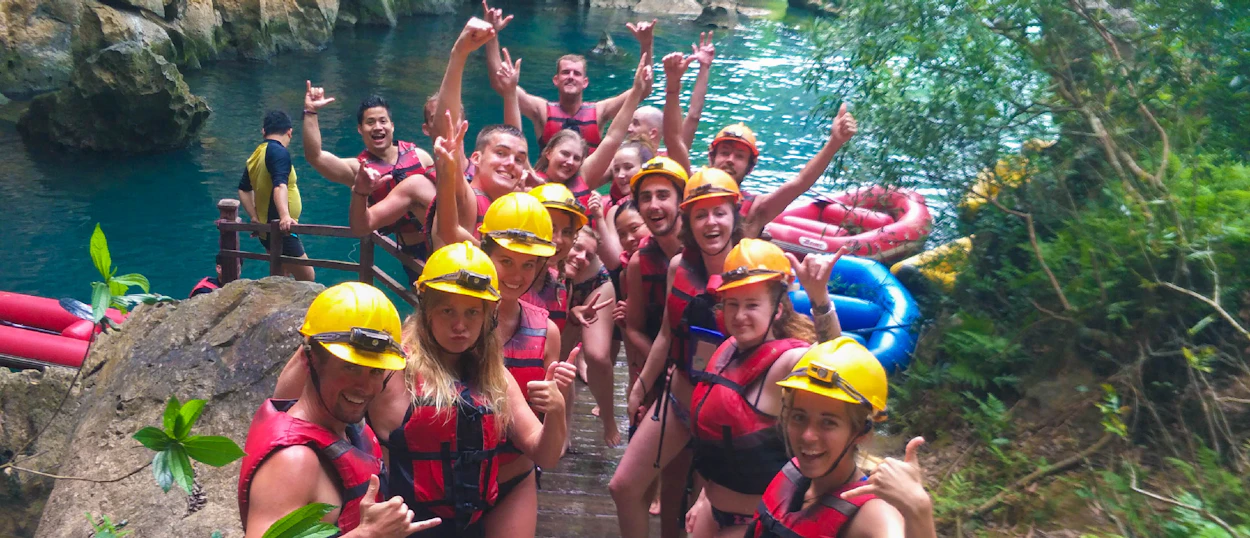 A large group of smiling adventurers in helmets and life vests pose by clear water in Phong Nha.