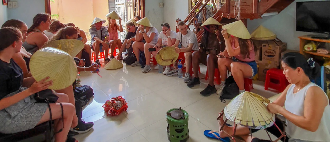 A group of travelers in Hue, Vietnam, learning to craft traditional conical hats from a local artisan.