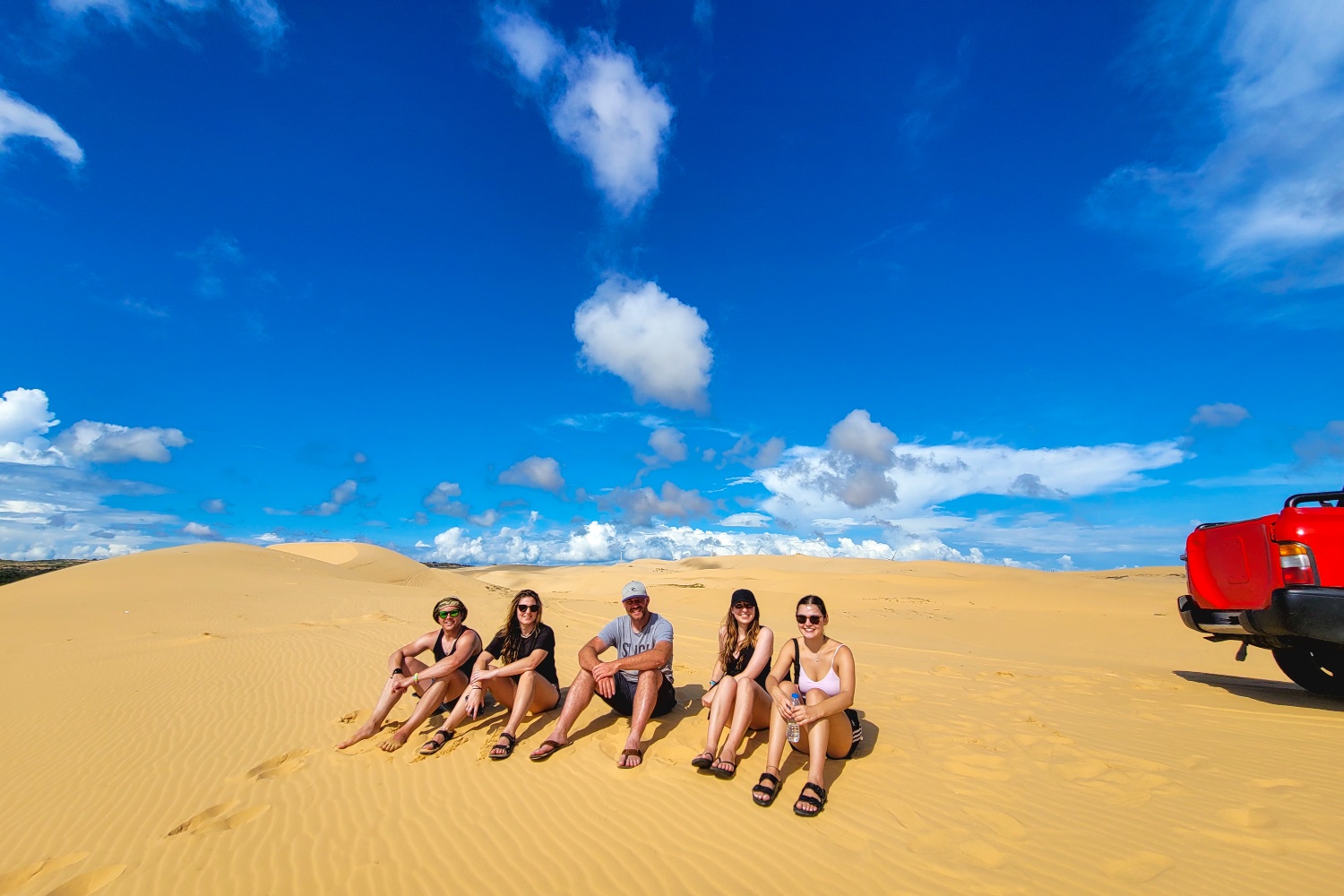 Five friends relax on golden sand dunes under a bright blue sky in Vietnam.