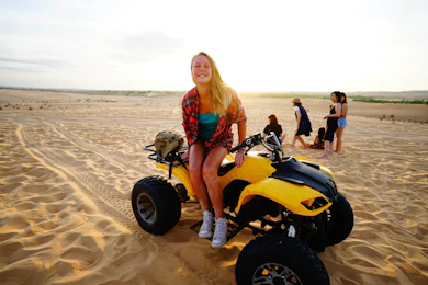 Blonde woman smiles from a yellow ATV on sandy dunes with other travelers in Vietnam.