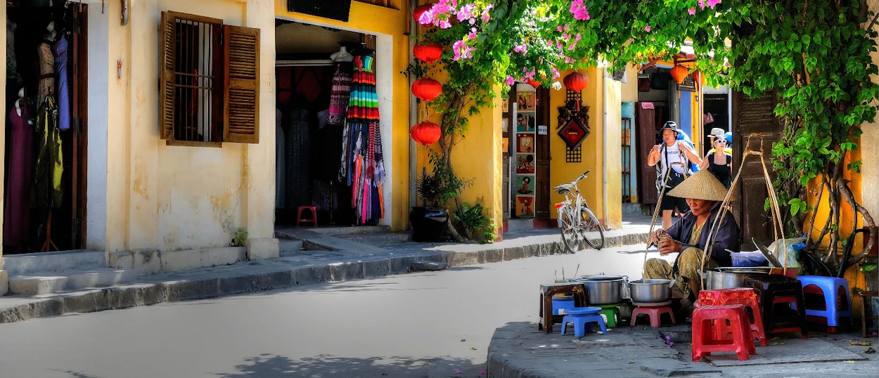 A person in a conical hat sells street food under flowering vines and red lanterns in Hoi An.