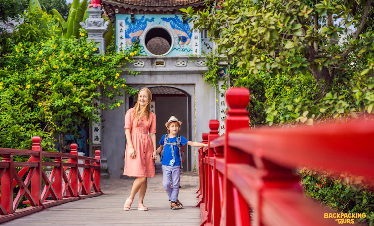 A woman and child walk on a red bridge towards a traditional temple gate in Hanoi.