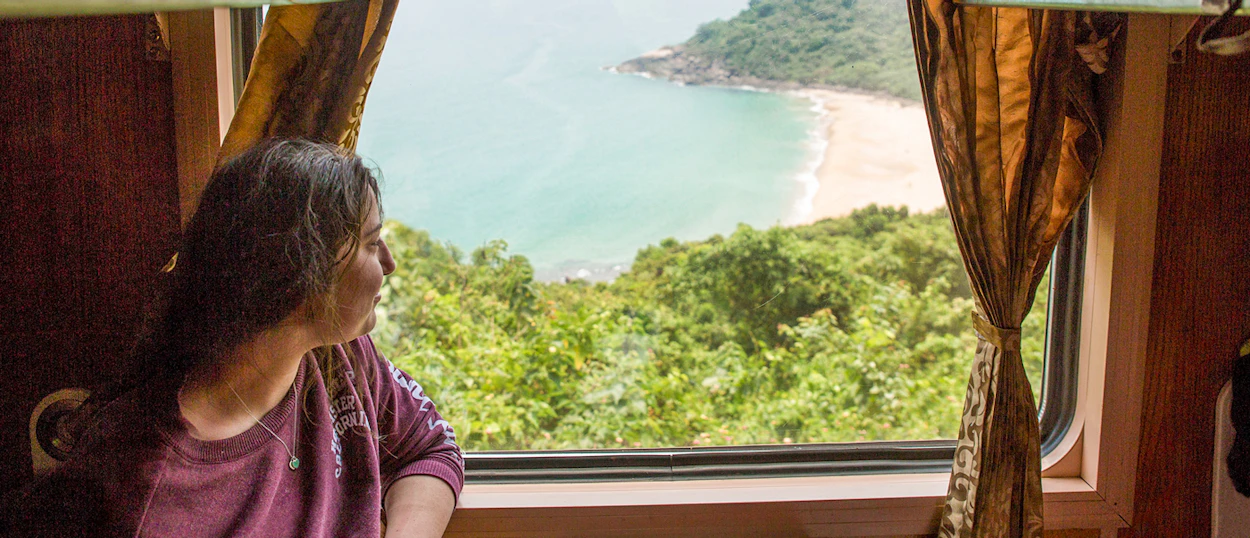 A woman looks out a train window at a beautiful sandy beach and turquoise ocean.