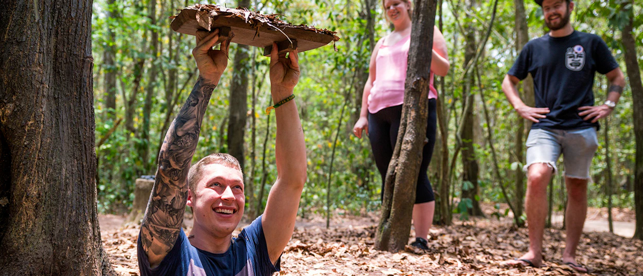 A smiling man emerges from a camouflaged tunnel entrance at the Cu Chi Tunnels in Vietnam.