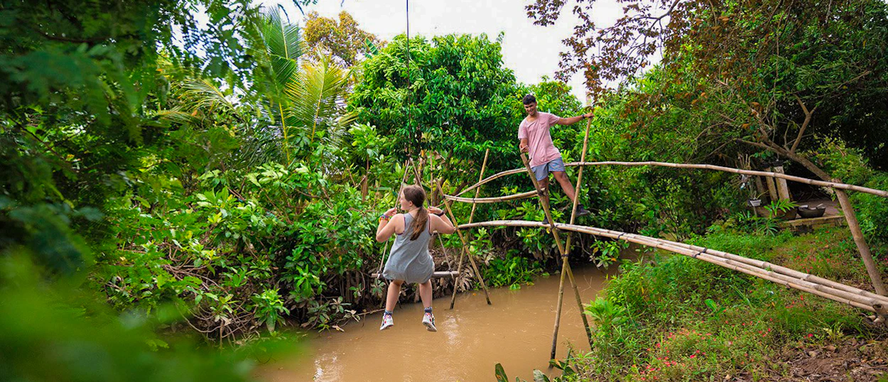 Adventurous travelers balance on a rickety bamboo bridge above a muddy river in the Mekong Delta.