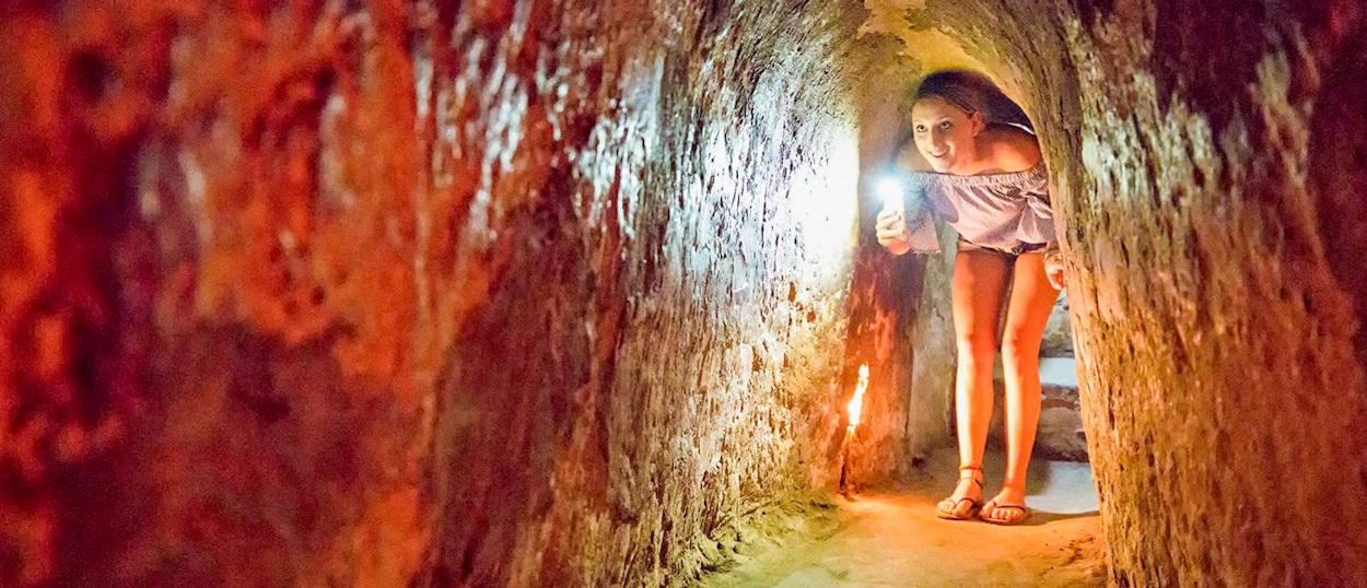 A young person smiles, holding a flashlight while exploring a narrow, reddish-brown tunnel at Cu Chi Tunnels.