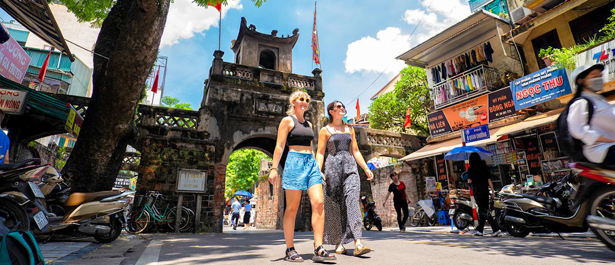 Smiling women walk past an ancient gate and busy shops on a sunny day in Hanoi.