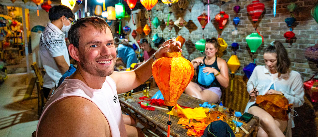 A smiling man holds his handmade orange lantern in a vibrant Hoi An workshop with other travelers.