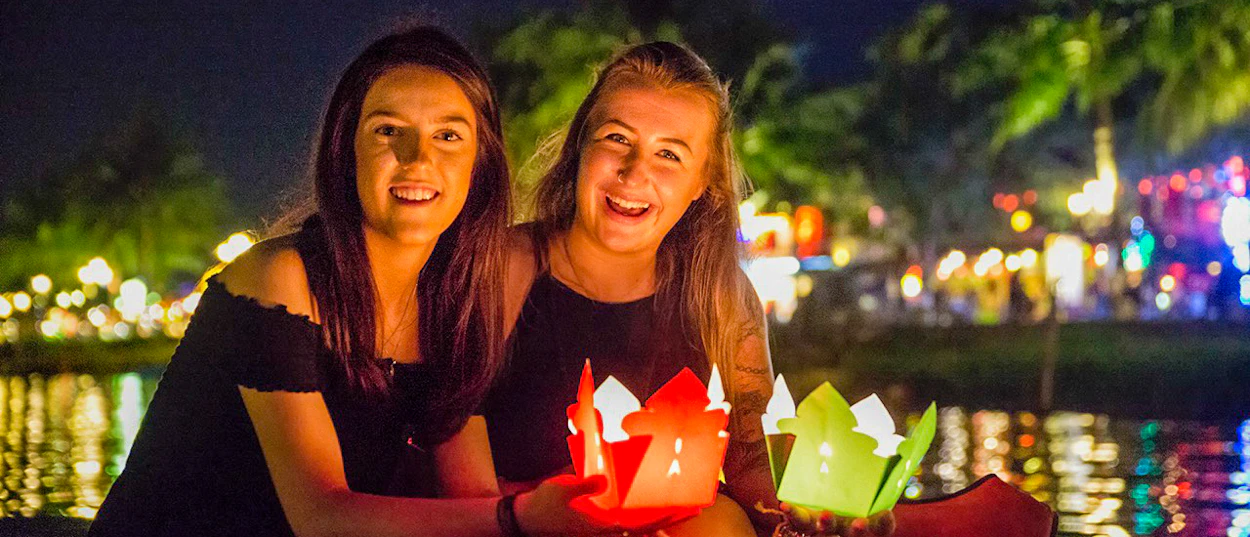 Smiling women holding colorful lanterns by the illuminated river in Hoi An at night.